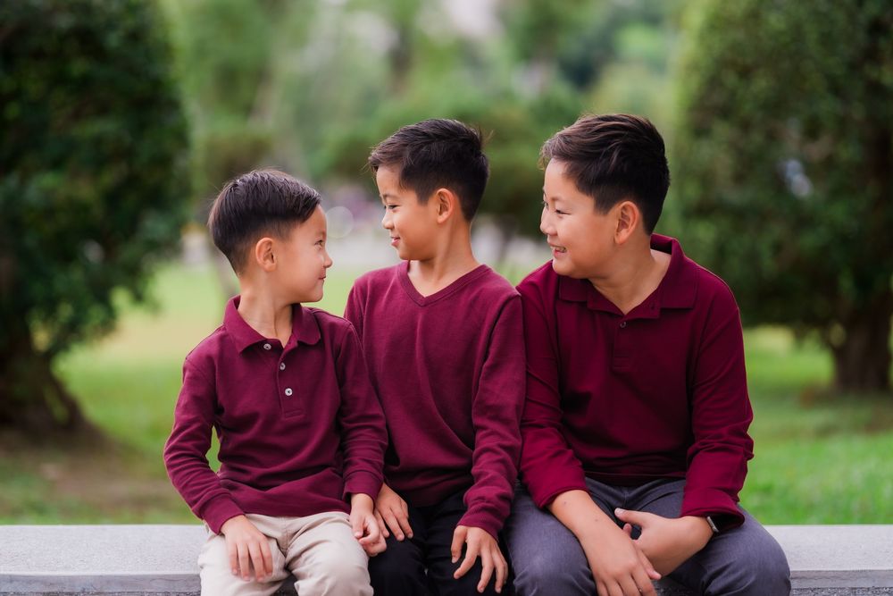 Three boys in matching burgundy sweaters share playful moments while sitting outdoors in a park setting.