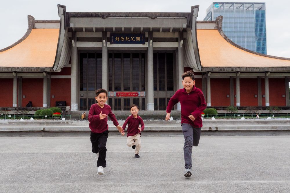 Two people run across a large plaza in front of a traditional Asian temple building with red pillars and curved roofs.