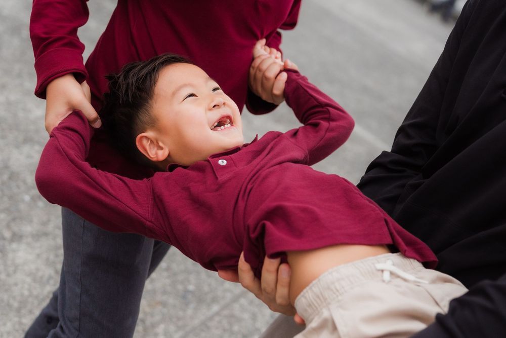 A child in a burgundy sweater laughs joyfully while being playfully swung by adults in a candid moment.