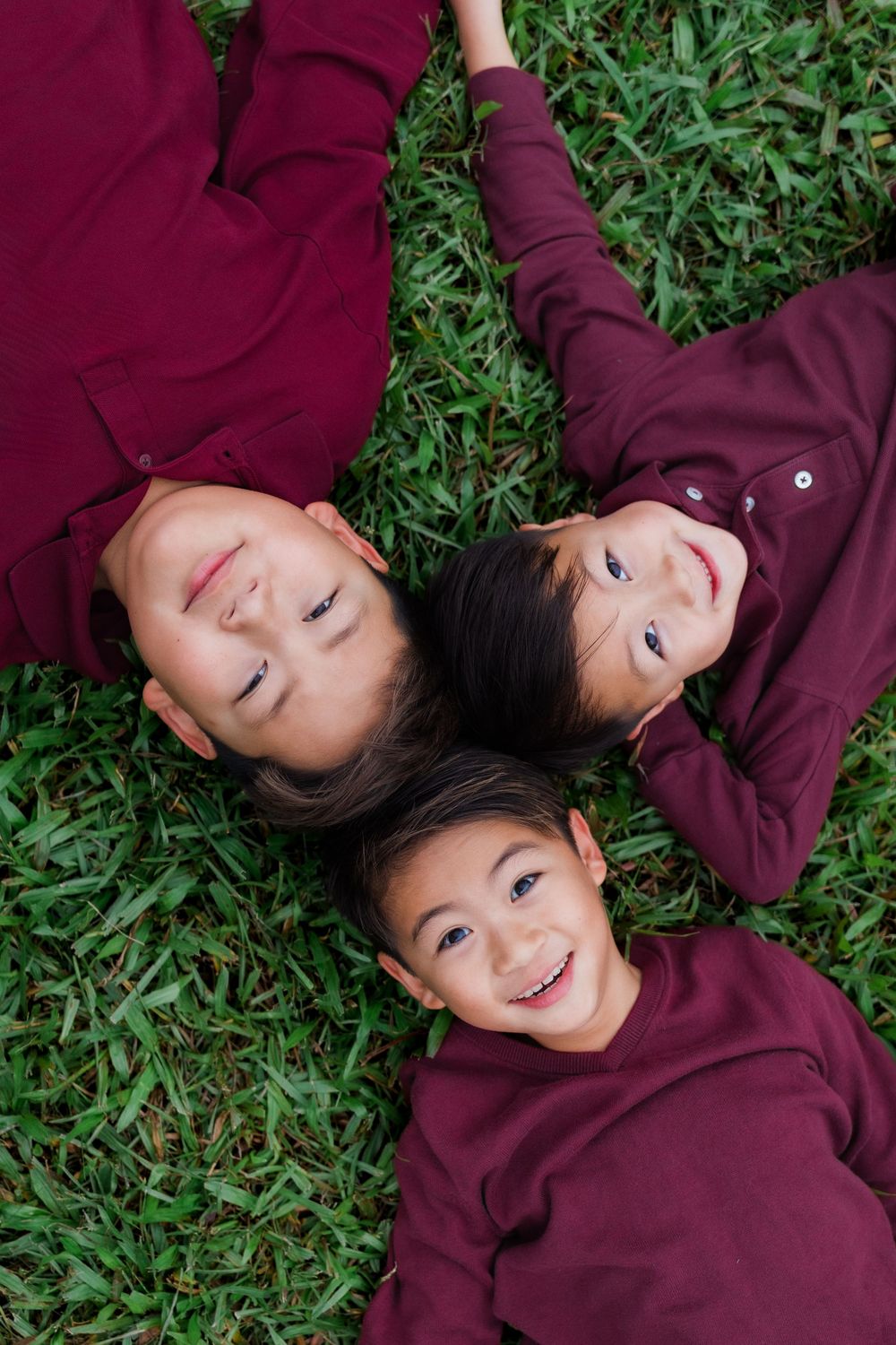 People in burgundy sweaters lie on green grass while smiling and laughing together.