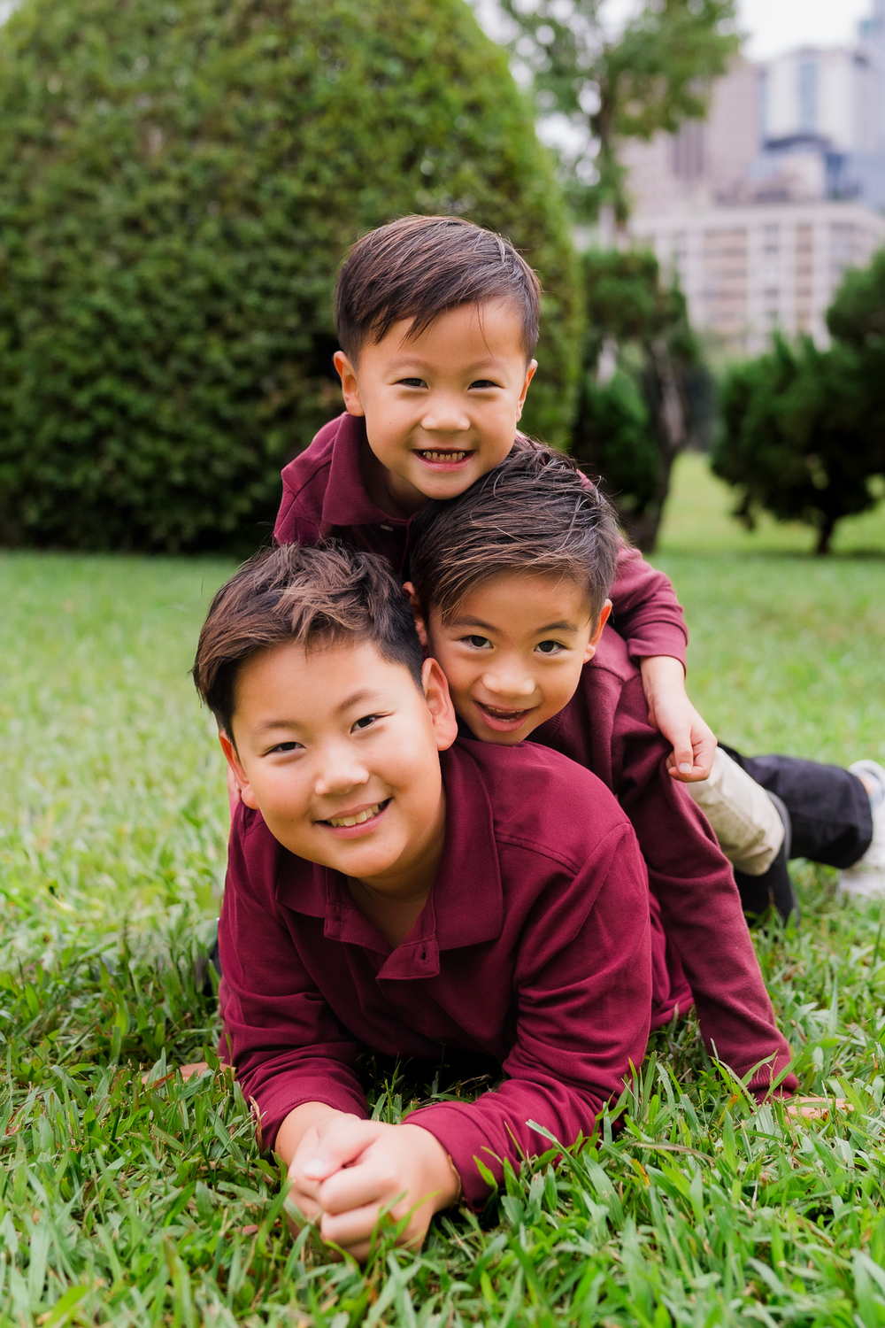 Two children in burgundy sweaters play and laugh together while lying in green grass on a sunny day.