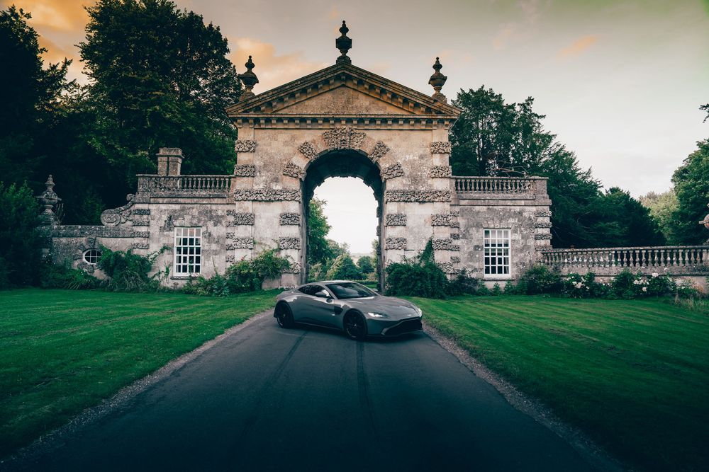 A grand stone mansion with classical architecture sits at the end of a long driveway surrounded by green lawns.