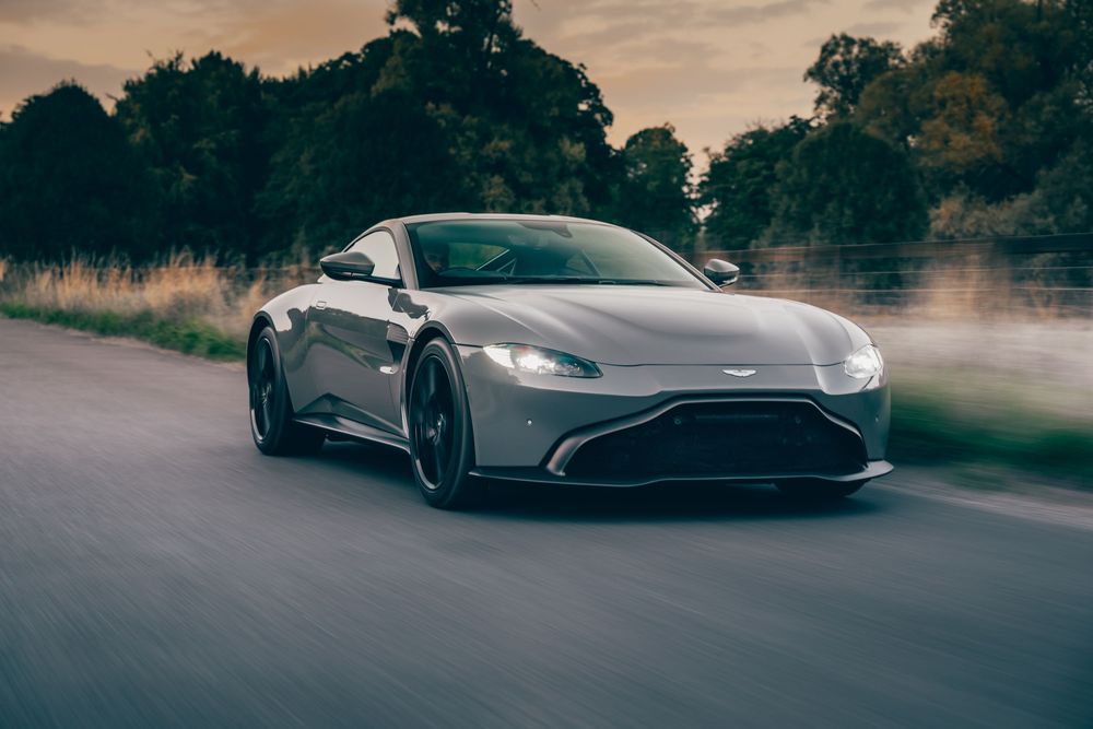 A sleek silver Aston Martin Vantage sports car speeds along a misty road at dusk with dramatic lighting.