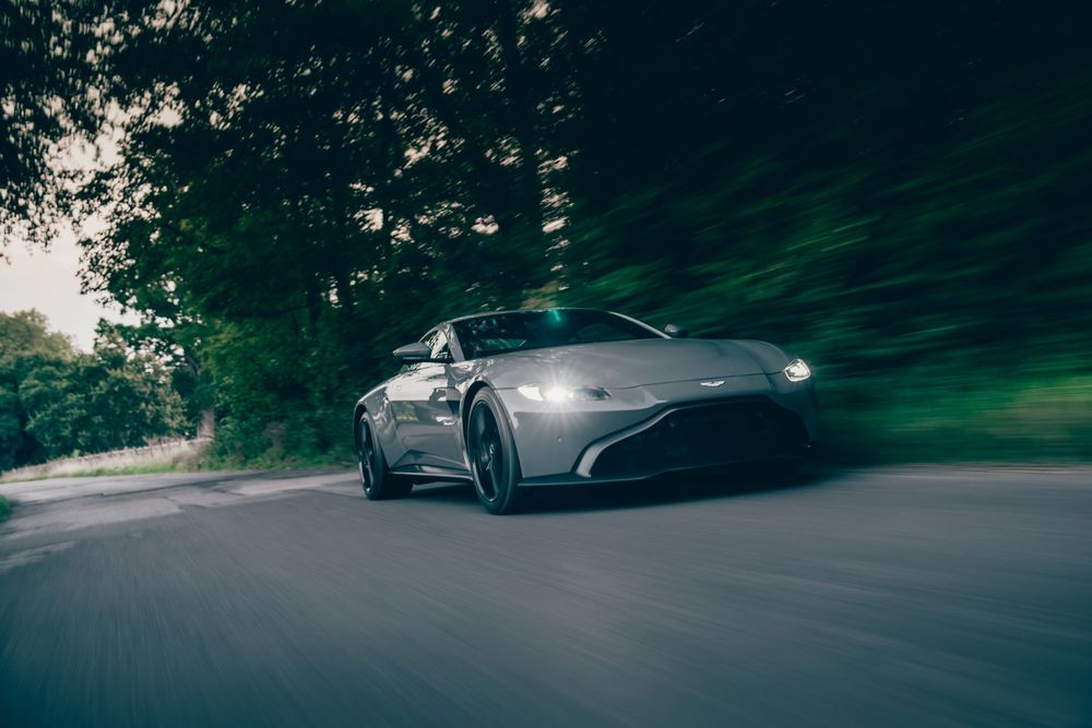 A sleek silver sports car drives through a lush green forest road during dusk.