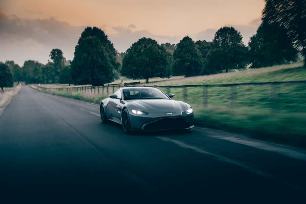 A black sports car drives down a tree-lined road during sunset creating a dramatic silhouette against the sky.