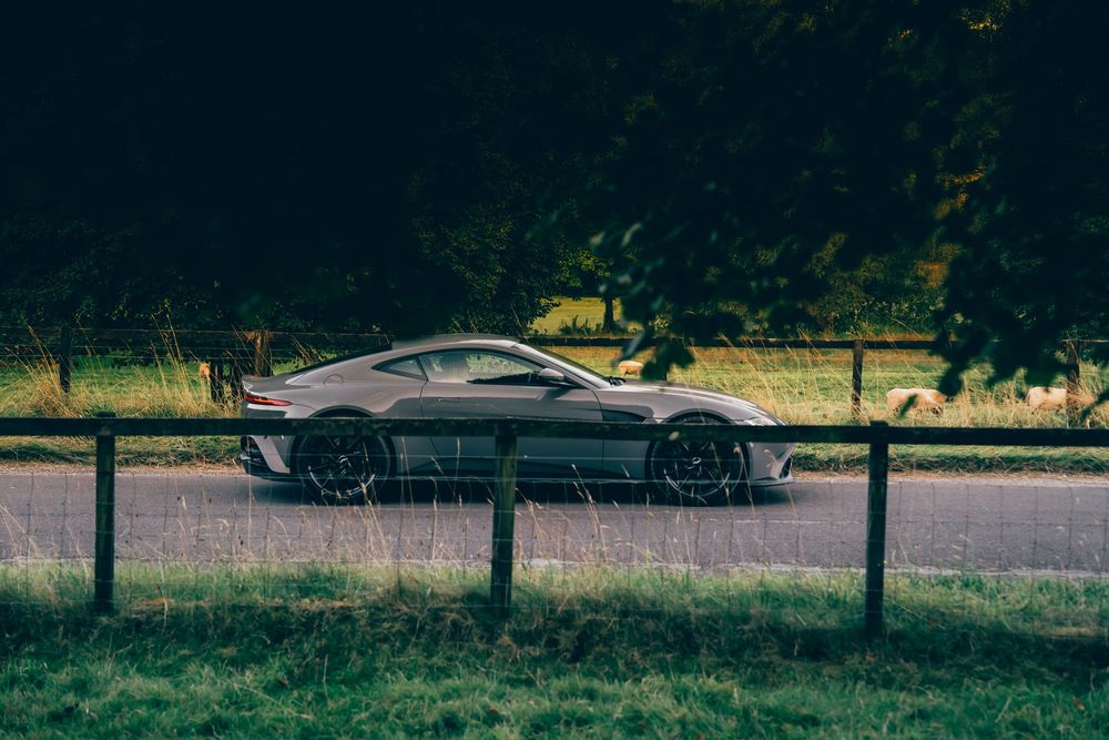 A silver sports car drives along a tree-lined country road on a sunny day.
