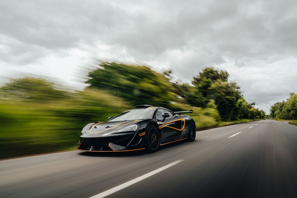 Black McLaren P1 supercar driving at speed on a winding country road under dramatic cloudy skies.