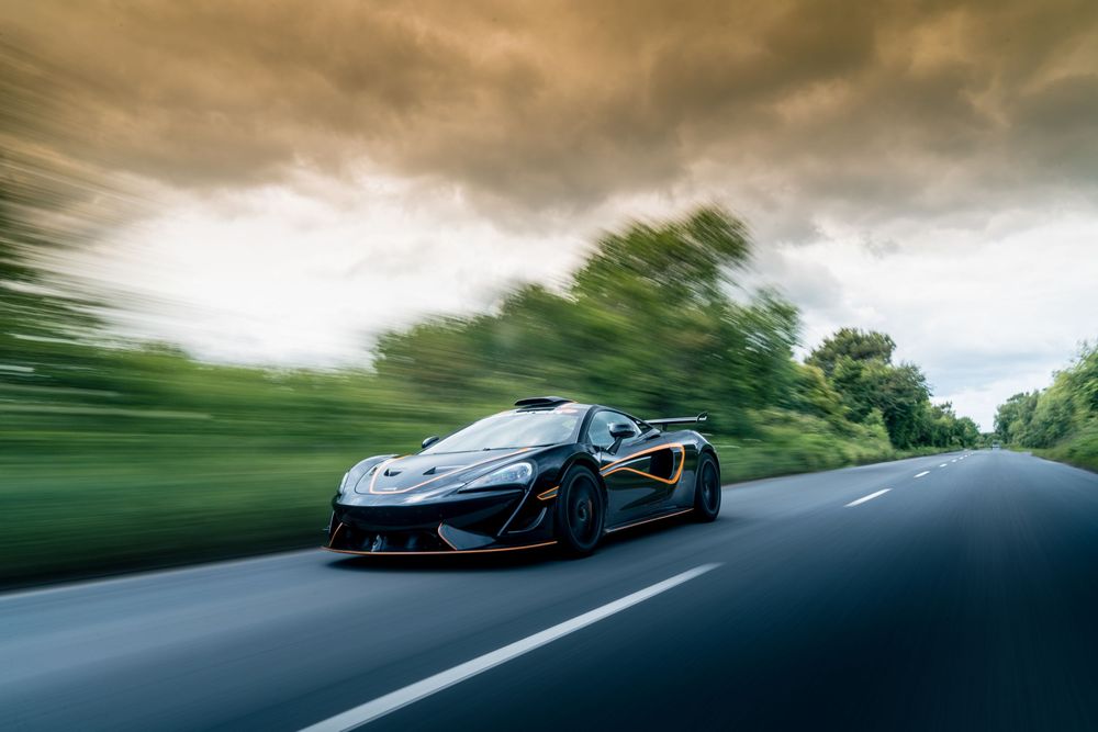 A McLaren supercar speeds along a winding road with dramatic cloudy skies overhead.