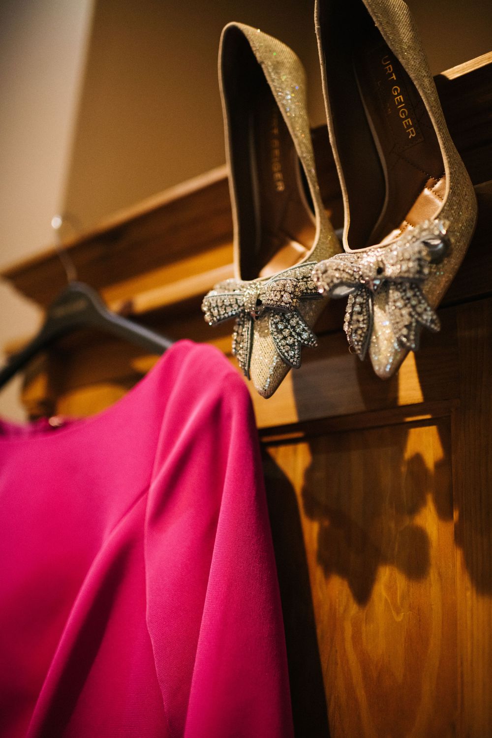 Elegant glittery high heels with crystal embellishments hanging on a wooden rack next to a pink garment.