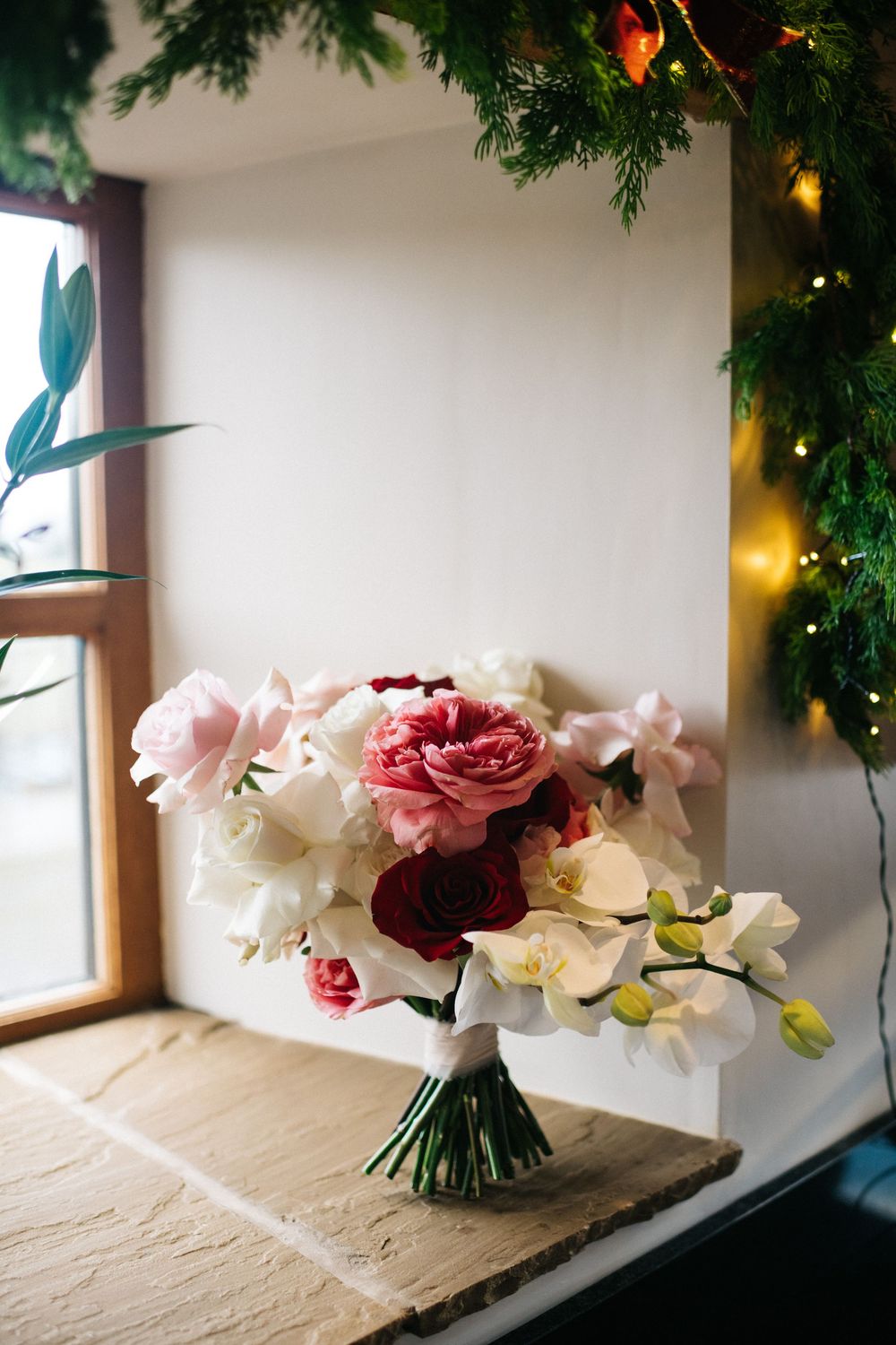 Elegant pink and white wedding bouquet with roses and orchids arranged on a rustic wooden surface with evergreen decor above.