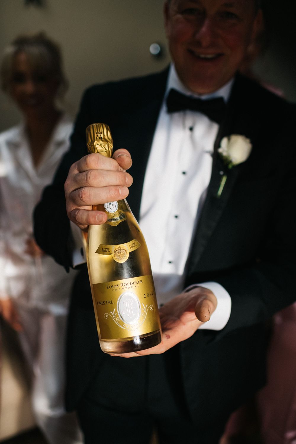 A person in formal attire holding a gold-labeled champagne bottle at what appears to be a celebratory event.