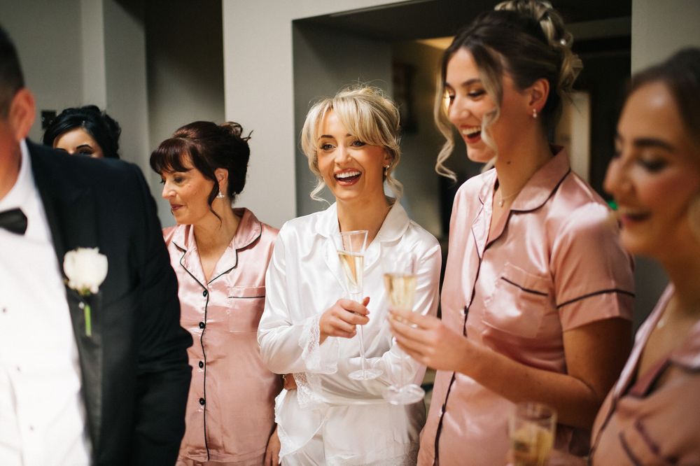 A group of bridesmaids in pink satin robes gather together laughing and smiling during wedding preparations.