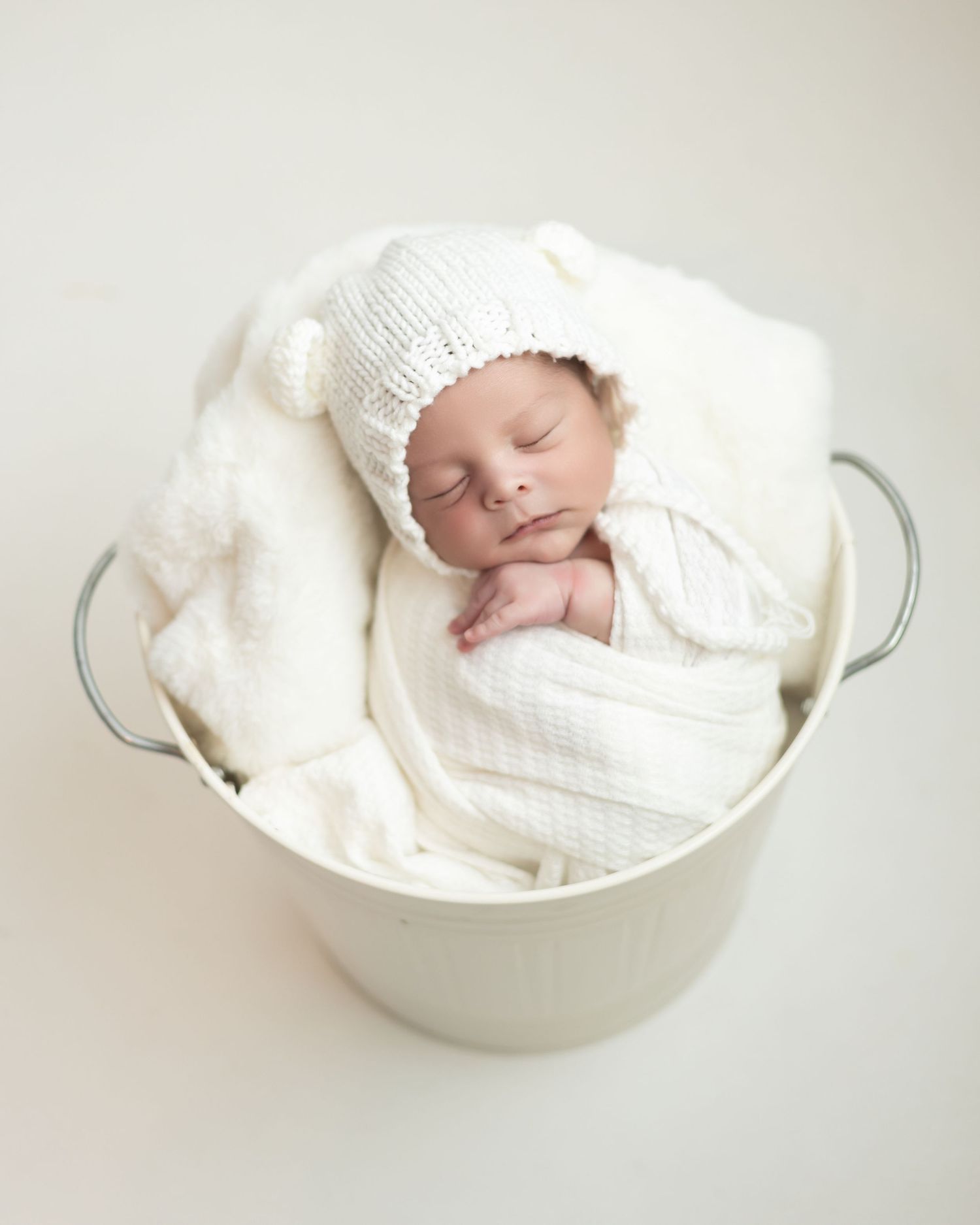 A sleeping newborn wrapped in white blankets rests peacefully in a metal bucket with soft white padding.