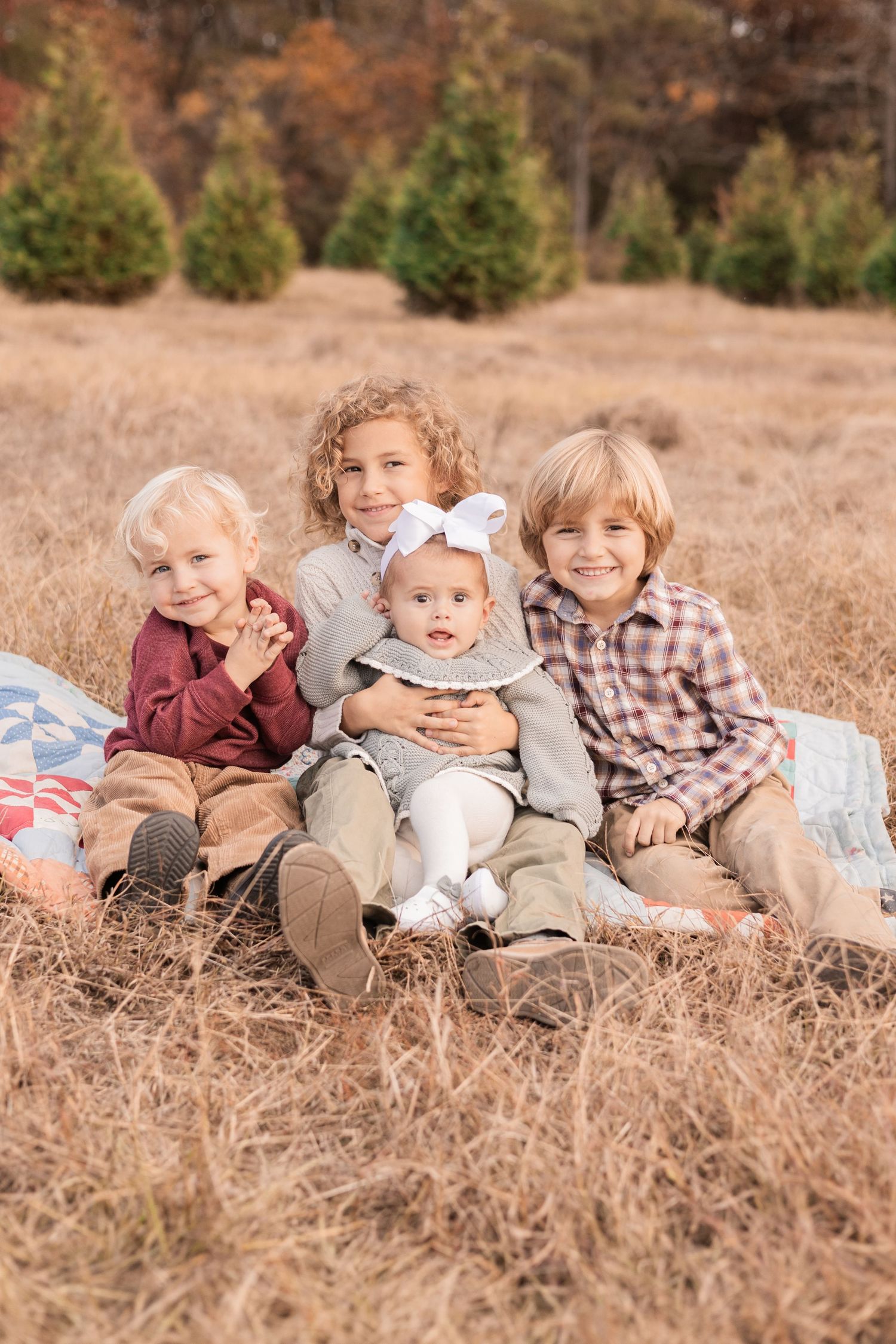 Children sitting together on a blanket in a brown autumn field with evergreen trees in the background in Pinehurst NC