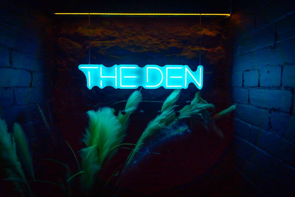 Neon blue sign reading 'THE DEN' illuminates a dark brick wall in a moody underground venue. Ellingham Hall