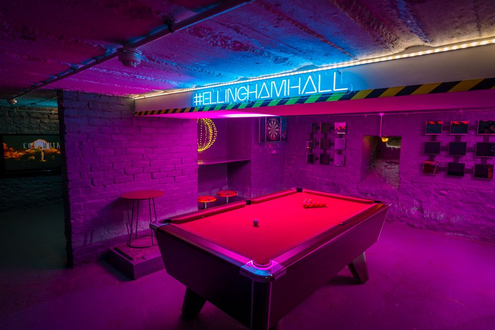 Pool tables and bar games illuminated by vibrant purple and blue neon lights in an underground venue. Ellingham Hall