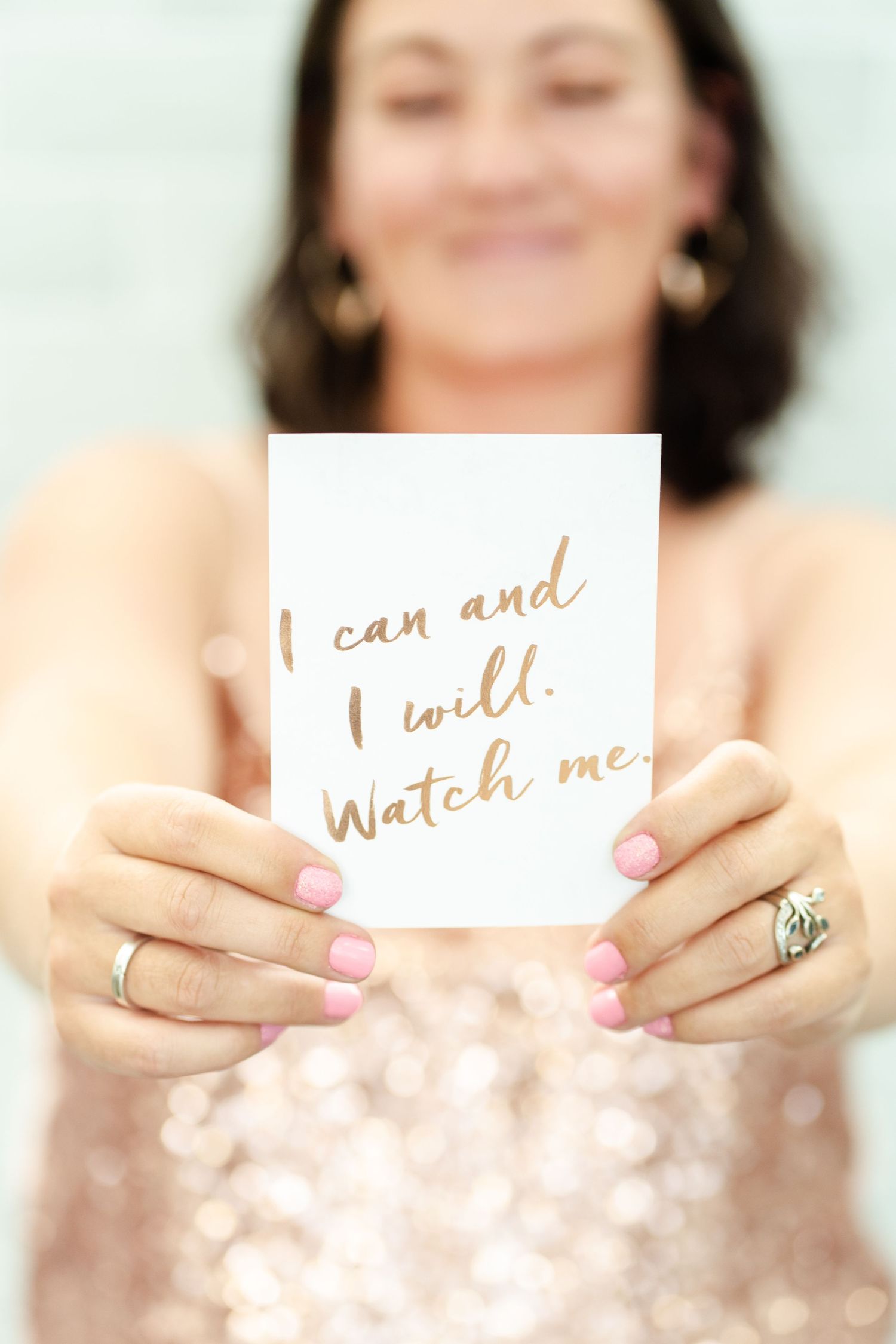 A woman smiles while holding a white paper note with a motivational phrase against a soft bokeh background.