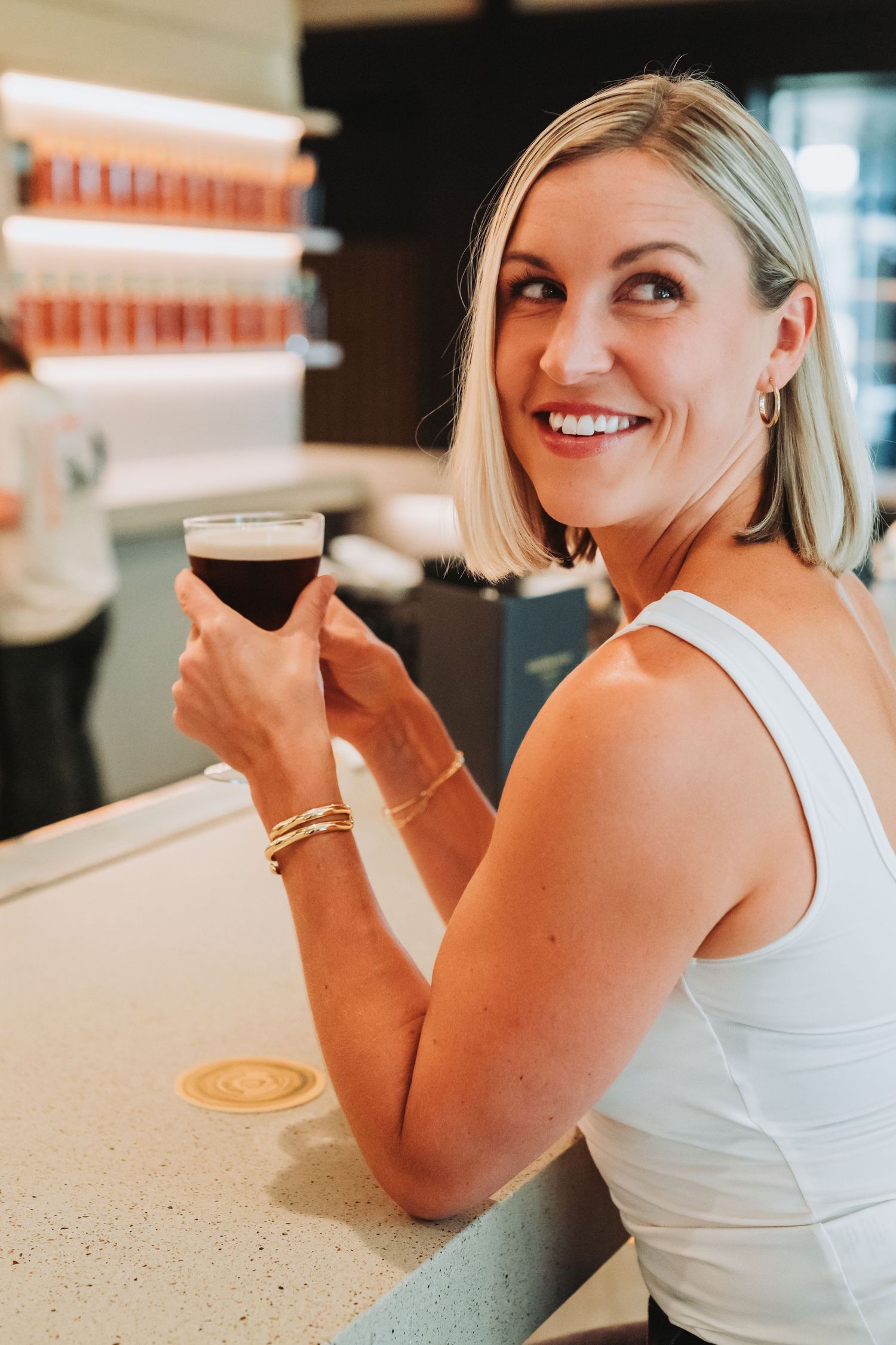 A person in a white tank top sits at a counter enjoying a frothy beverage with a big smile.
