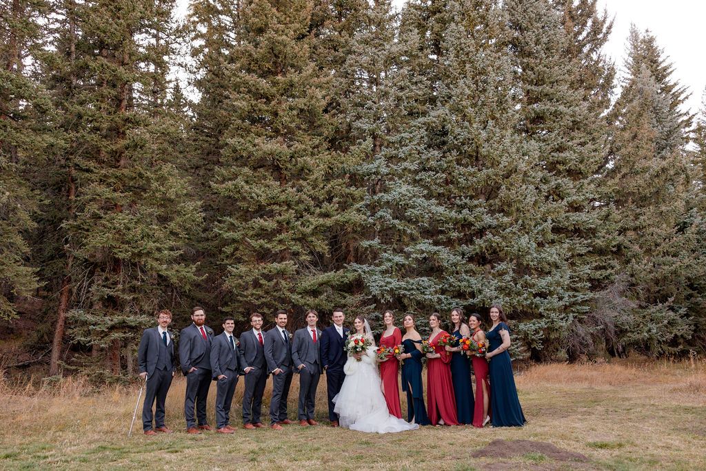 Bridal party in navy blue and burgundy dresses poses together in a rustic outdoor setting with evergreen trees.