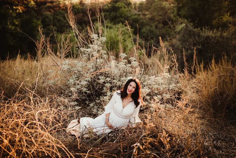 A person in a white dress sits in a field of golden dried grass and wildflowers during sunset.