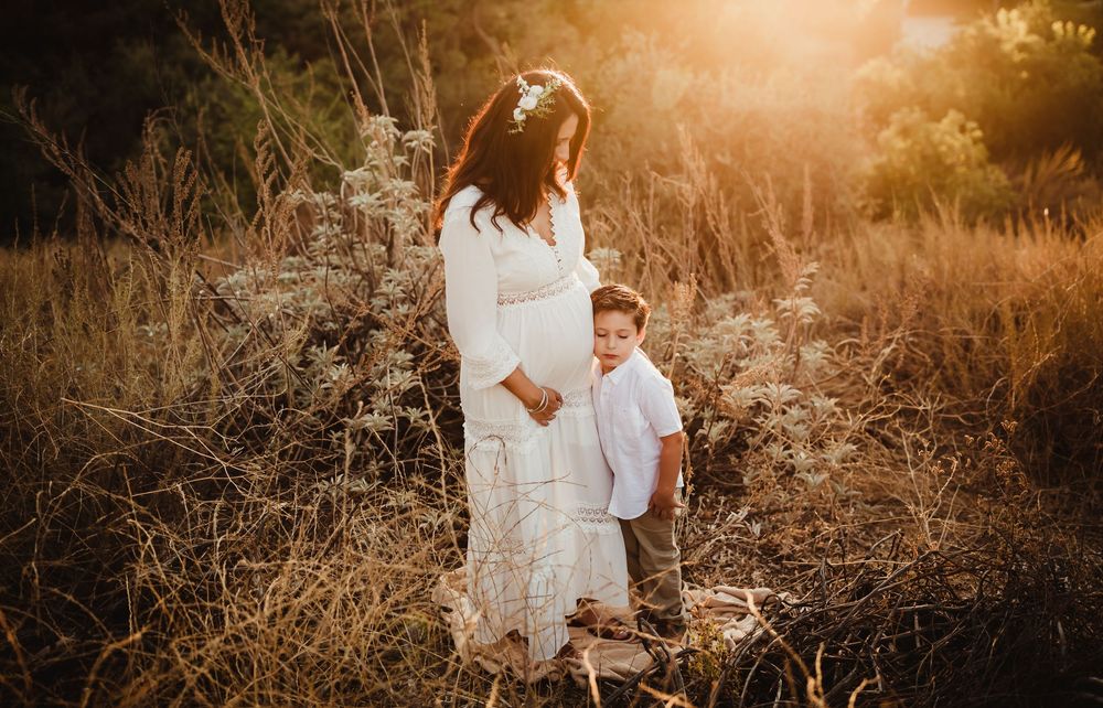 Ethereal golden sunset maternity photoshoot in tall grass field with white flowing dress creates dreamy atmosphere.