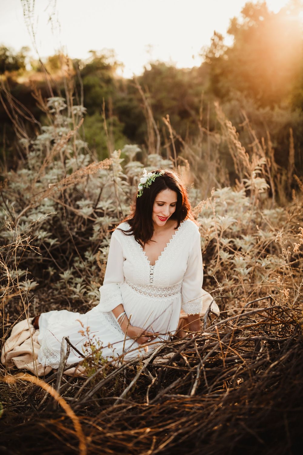A dreamy outdoor portrait session at golden hour with a person in a white lace dress sitting among dried grasses.
