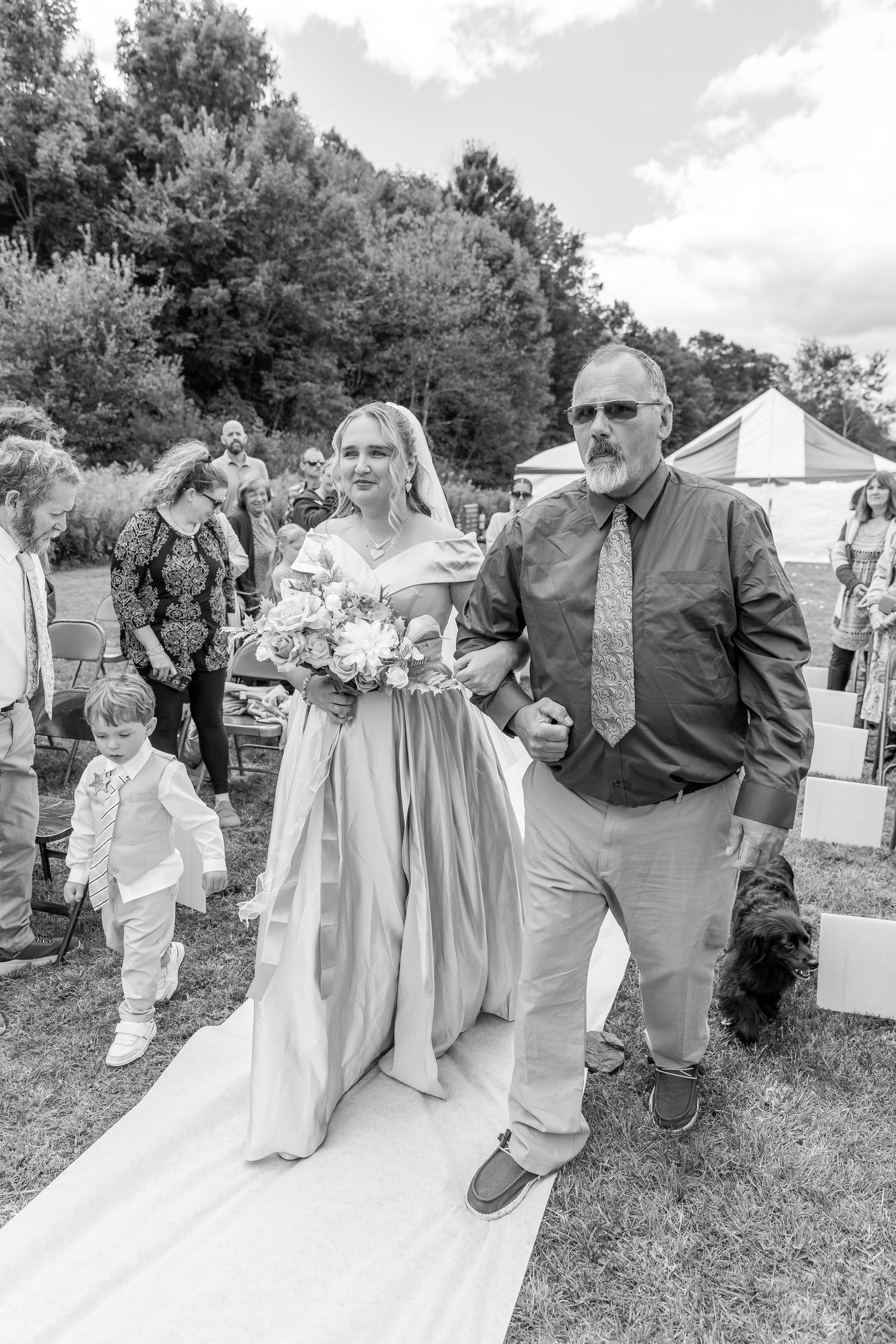 Bride in teal gown walks down outdoor aisle with older man during wedding ceremony, black and white.