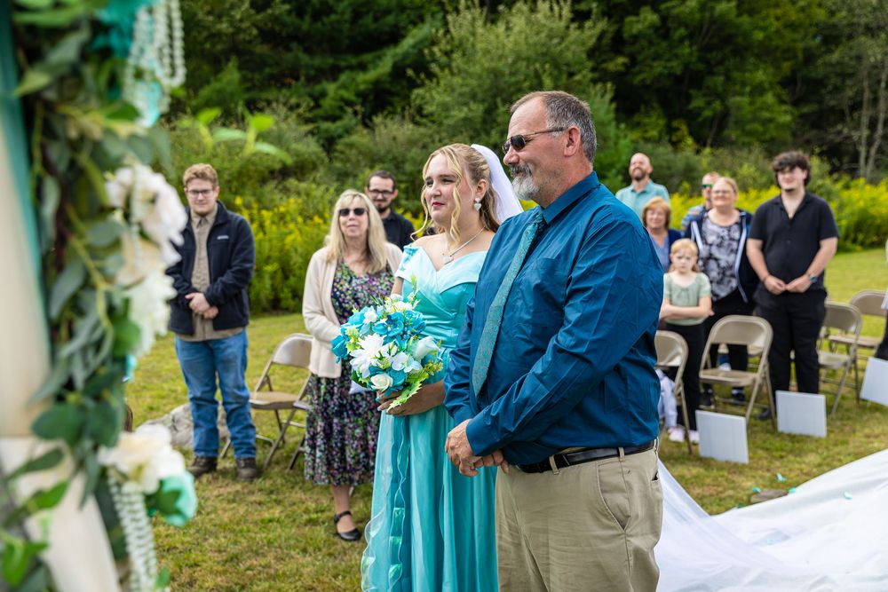 Smiling bride in teal dress holds bouquet while walking with escort at outdoor wedding ceremony.