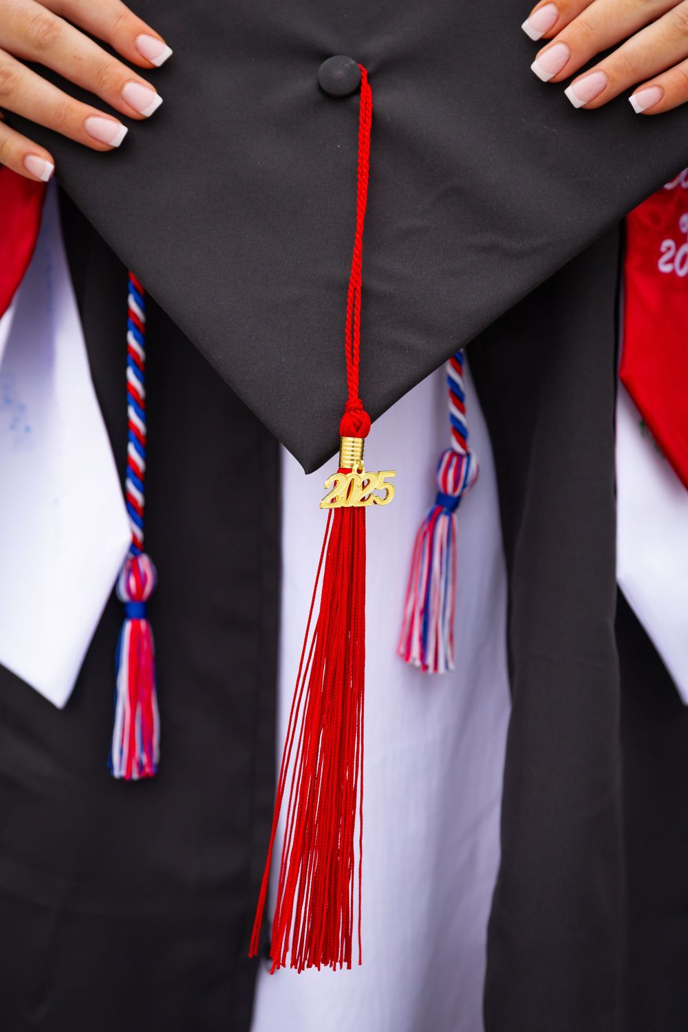 Close-up of graduation cap with gold 2025 charm and red tassel held by graduate in white gown.