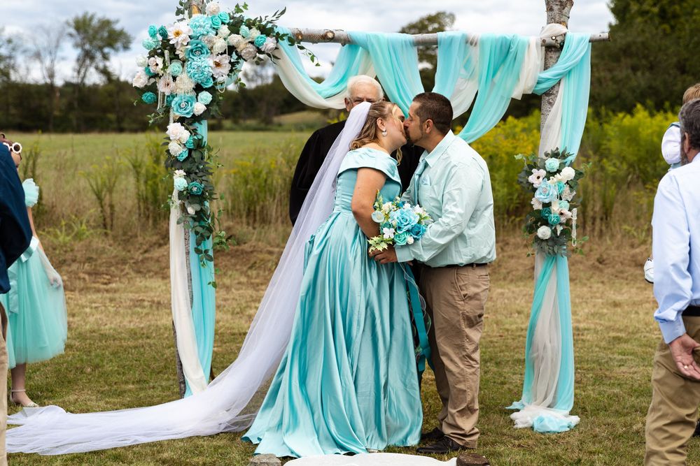 Bride and groom share first kiss under floral arch with teal decorations at outdoor wedding.