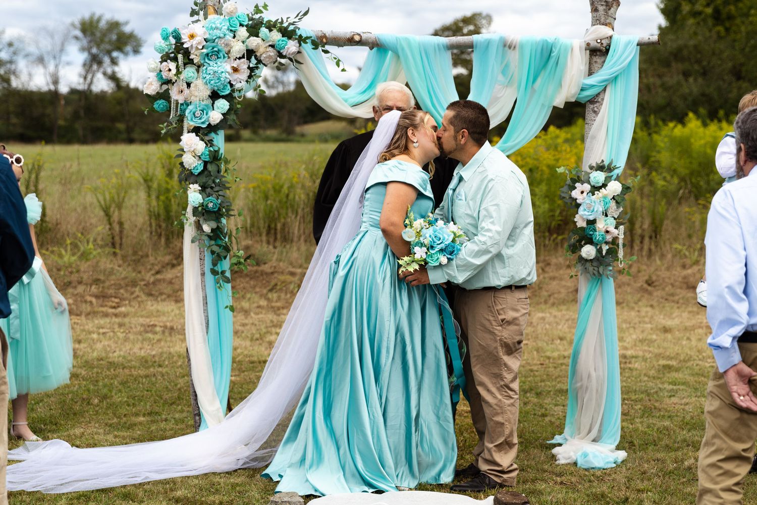 Bride and groom share first kiss under floral arch with teal decorations at outdoor wedding.