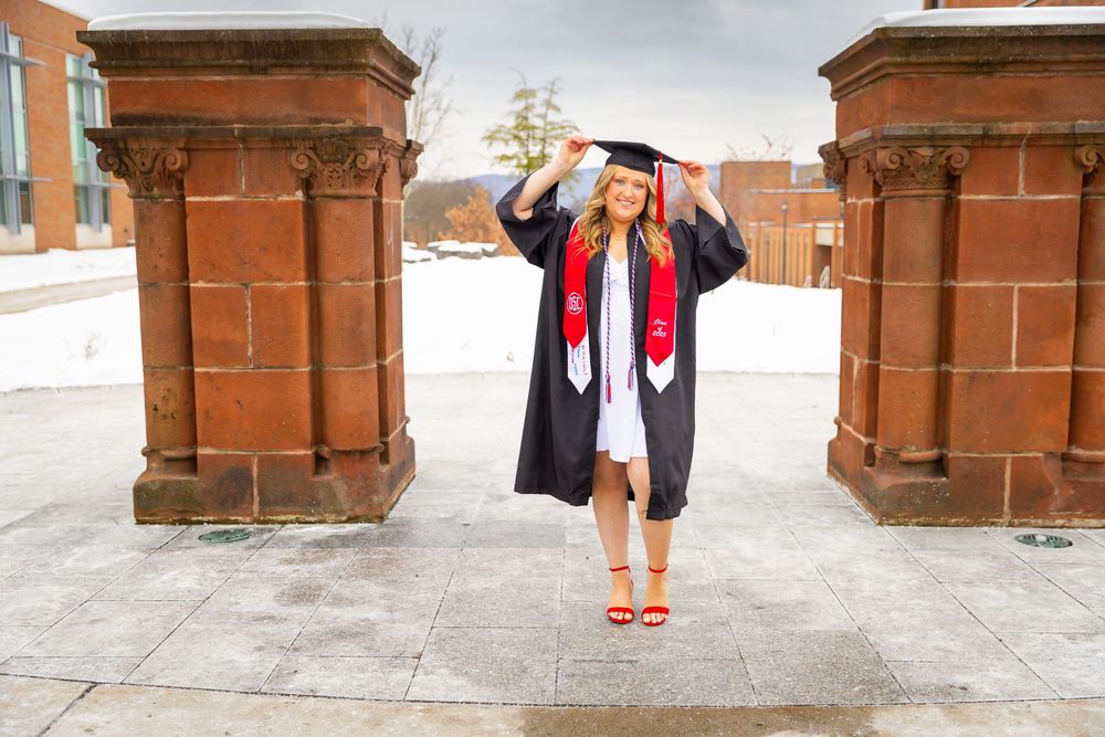 Female graduate in cap and gown posing between stone pillars at a snowy university campus.