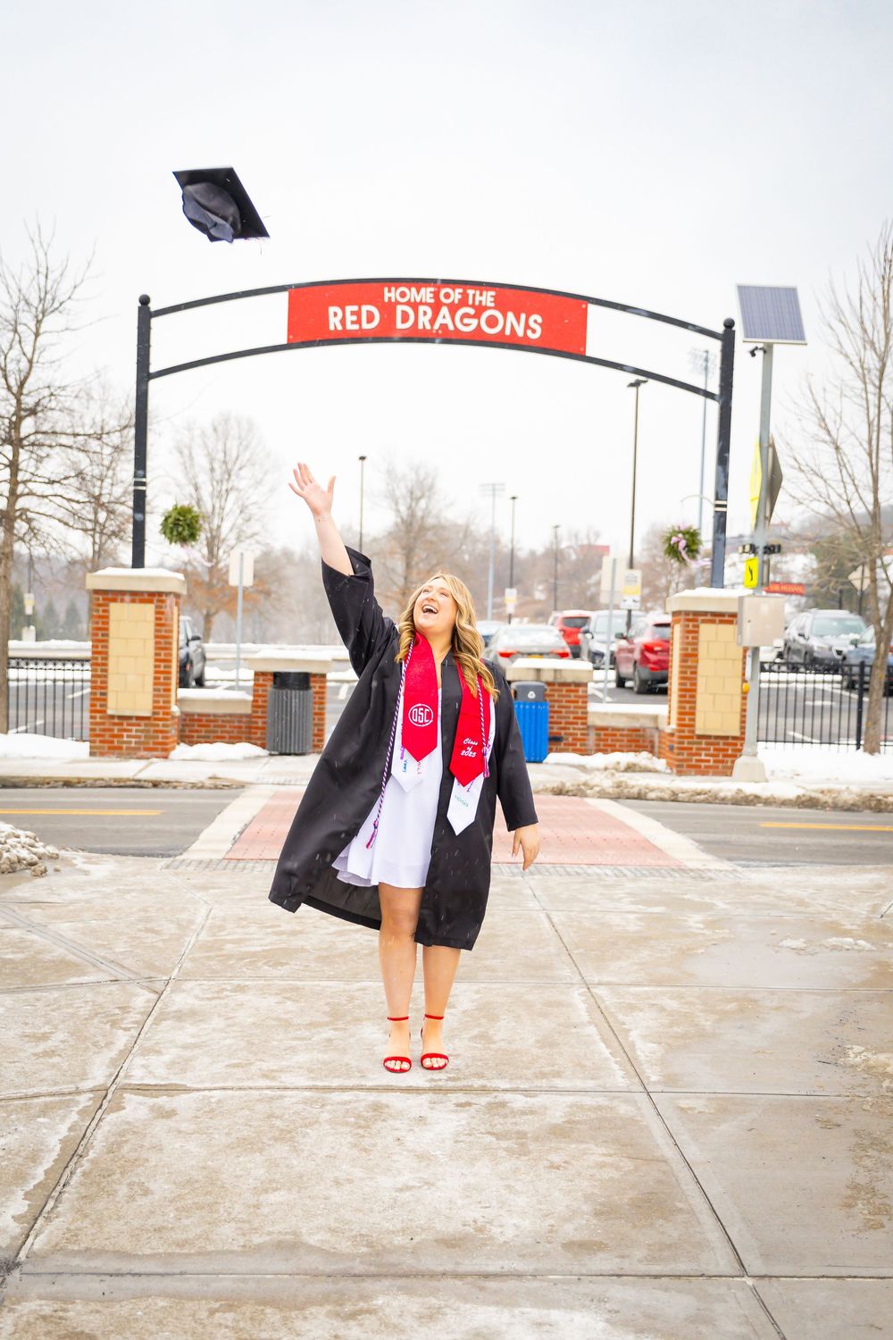 Female graduate tossing her cap in the air under a Red Dragons arch sign in winter.