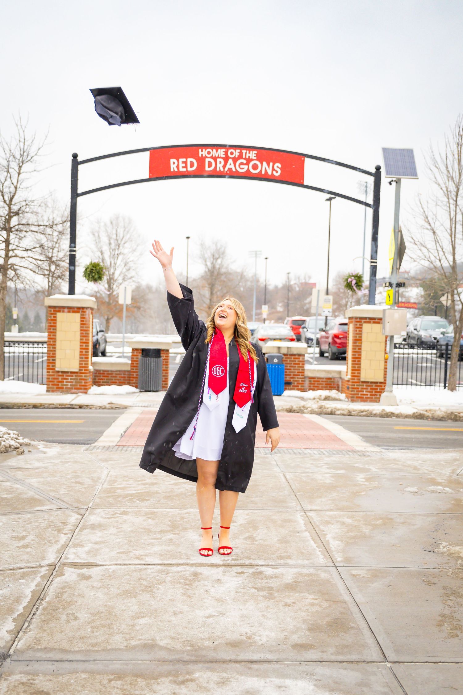 Female graduate tossing her cap in the air under a Red Dragons arch sign in winter.