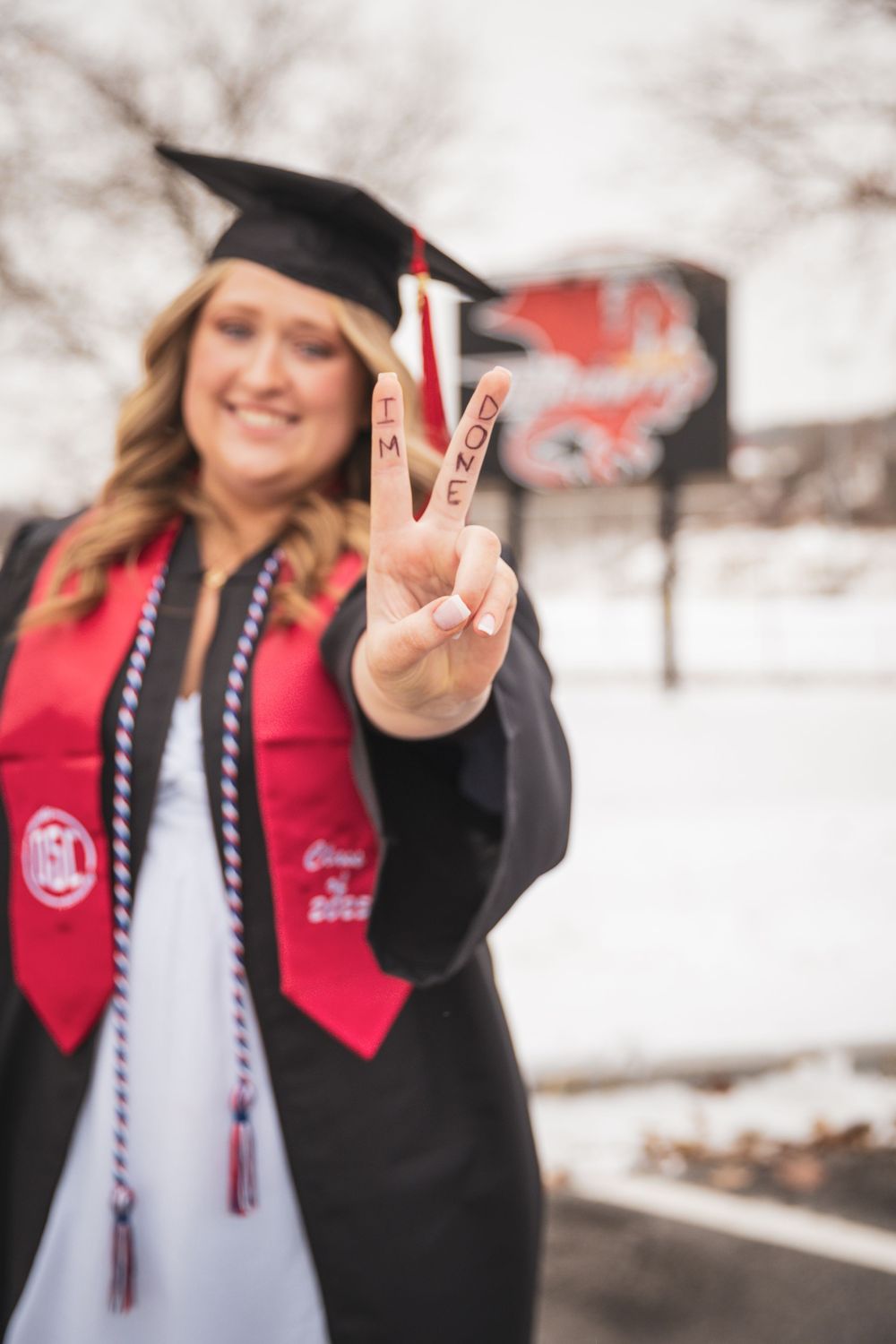 Smiling female graduate in cap and gown showing a peace sign with fingers at a snowy campus.