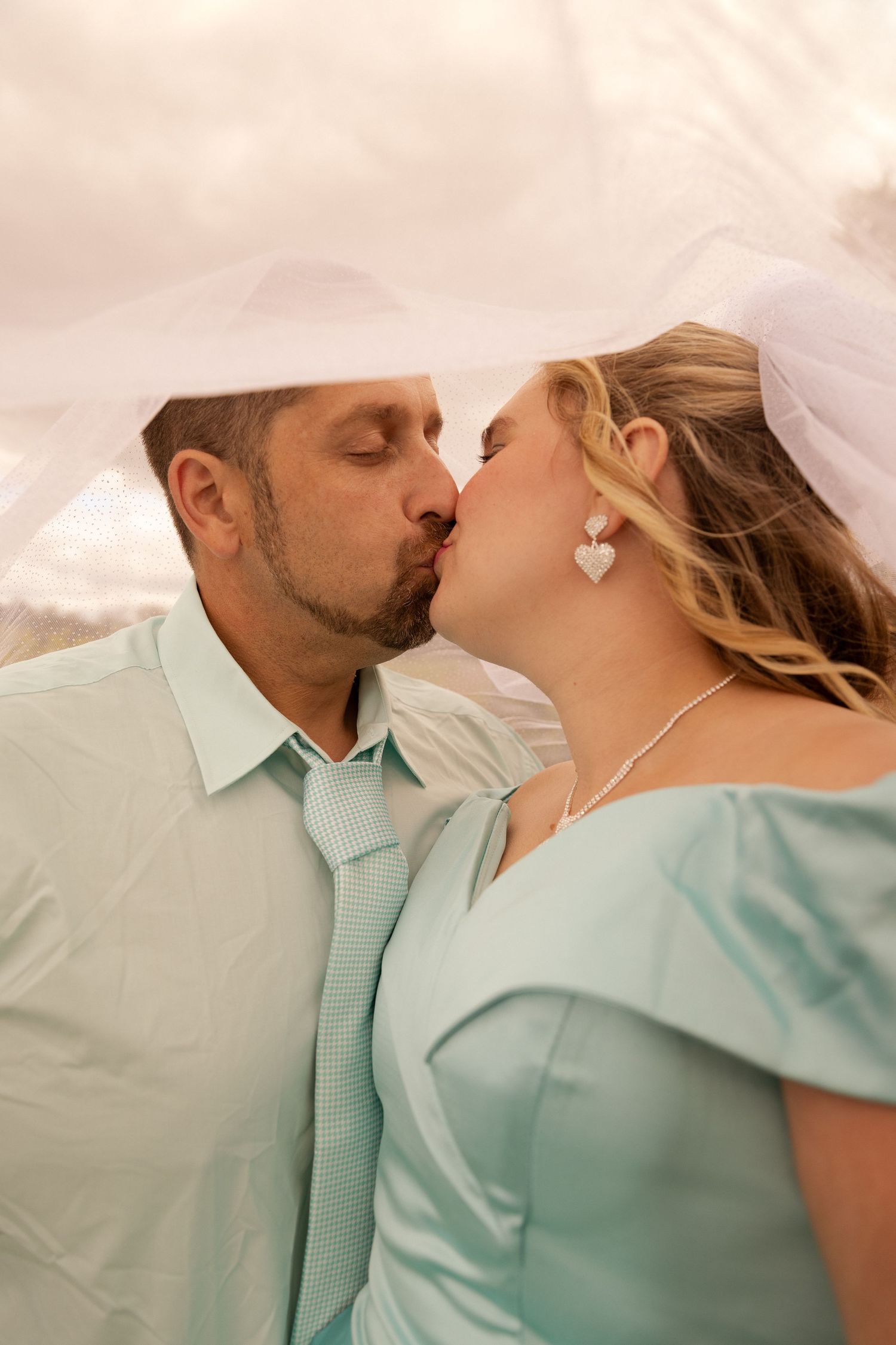 Bride and groom share romantic kiss under flowing white veil at outdoor wedding ceremony.