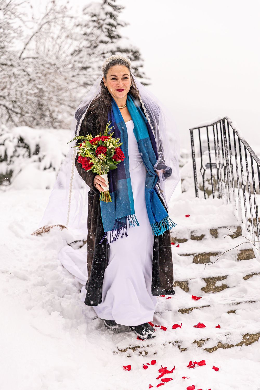 Winter bride in white gown, blue scarf and fur coat holding red rose bouquet on snow-covered steps.