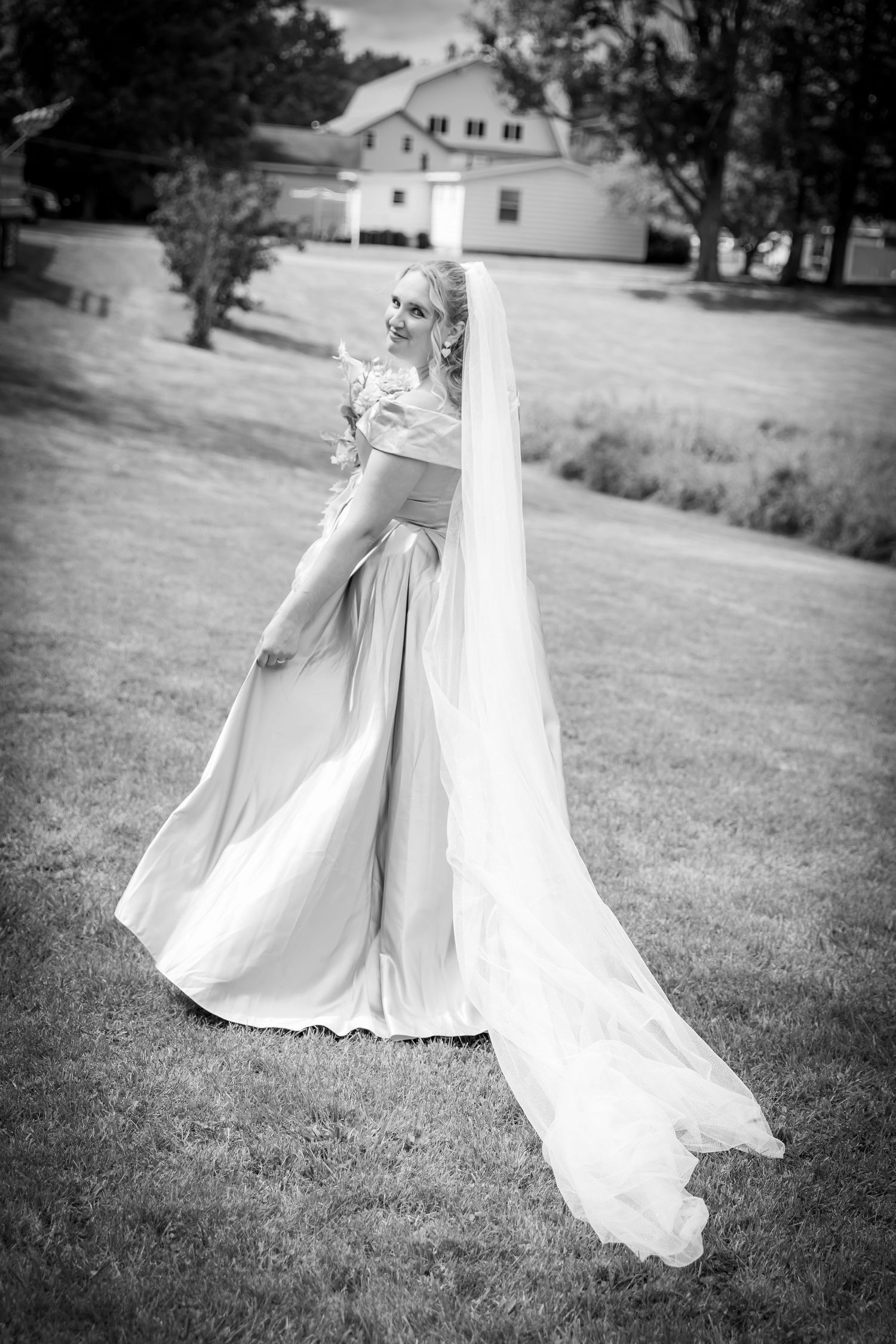 Elegant bride in flowing gown and long veil poses on green lawn near white farmhouse, black and white.