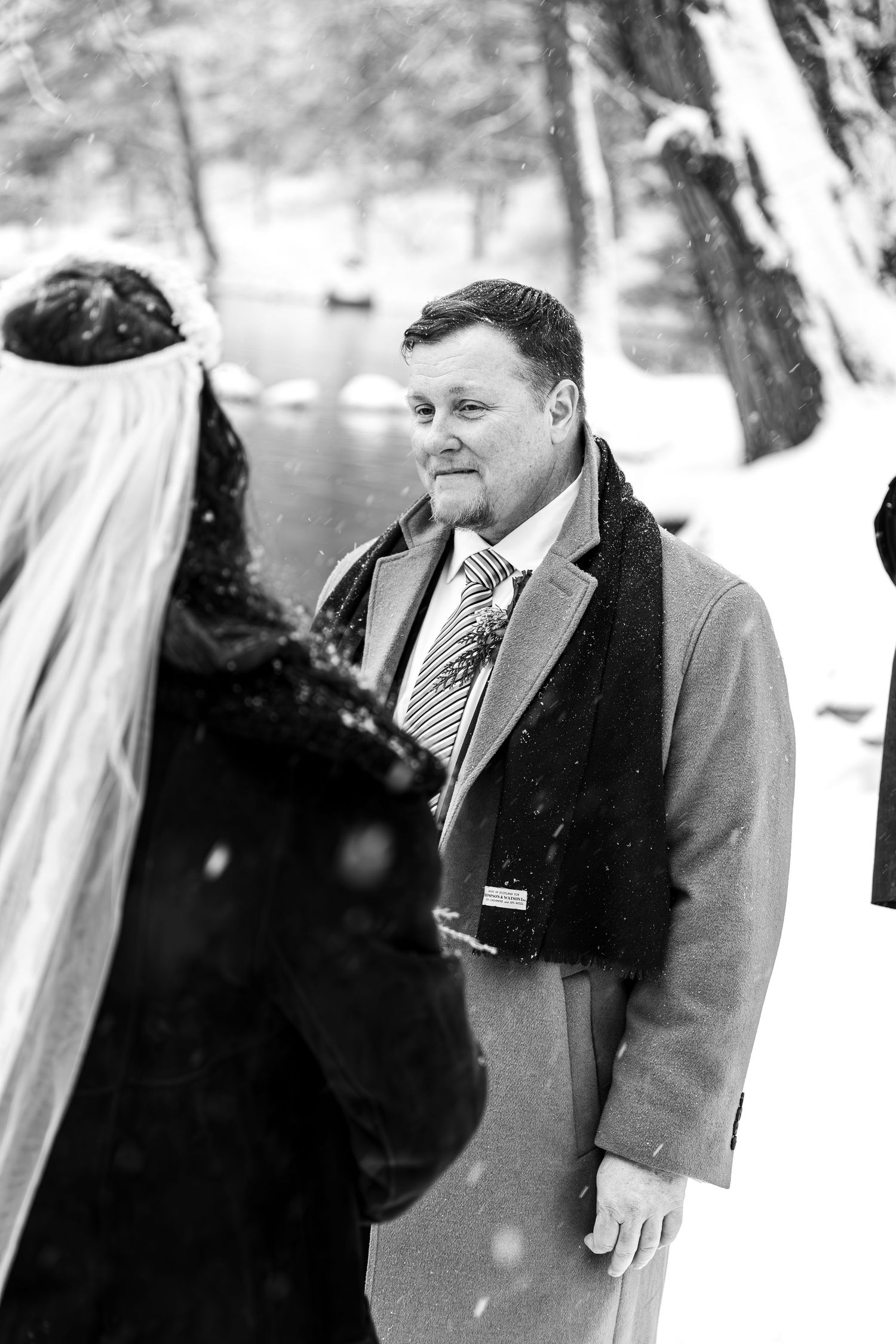 Black and white photo of groom in overcoat smiling at bride during outdoor winter wedding ceremony.