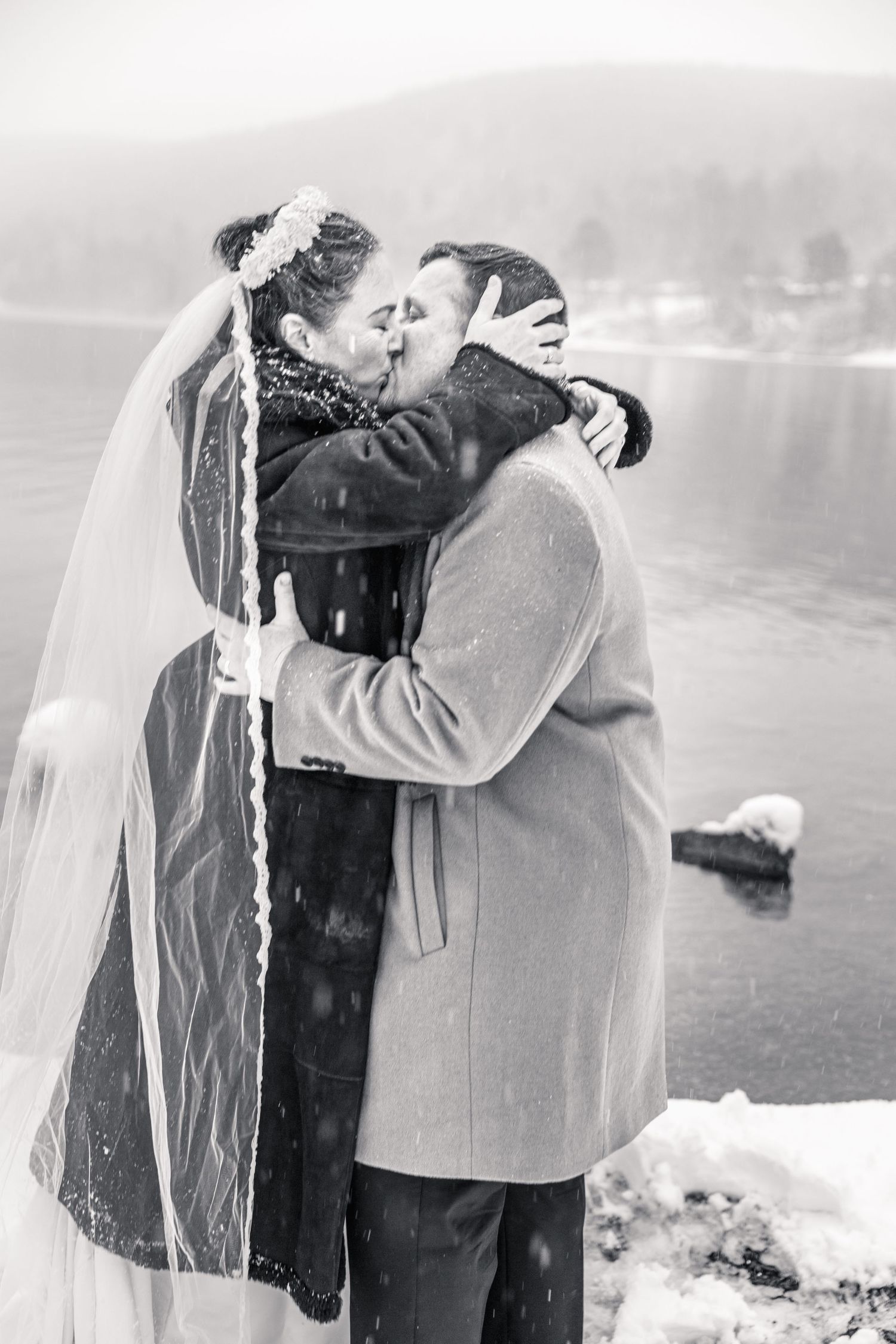 Black and white photo of newlywed couple sharing first kiss by snowy lakeside during winter wedding.