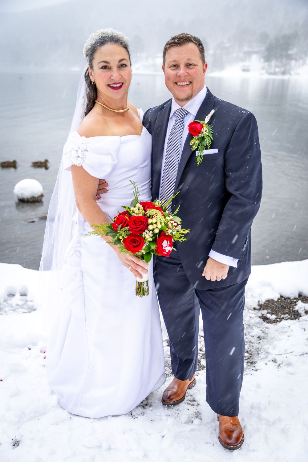Smiling bride and groom posing with red rose bouquet beside a snowy lake during winter wedding.