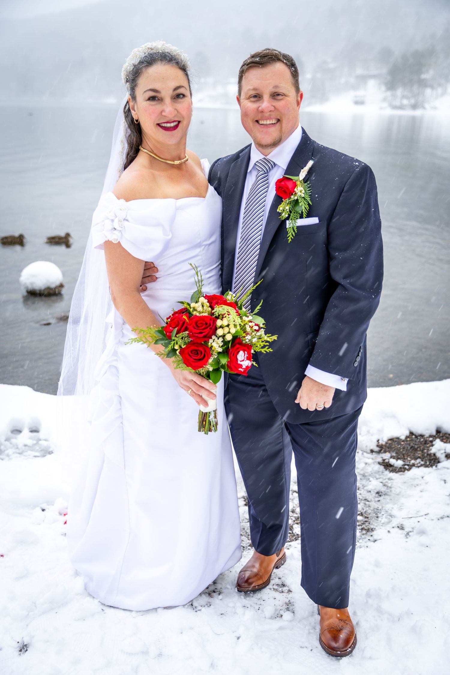 Smiling bride and groom posing with red rose bouquet beside a snowy lake during winter wedding.