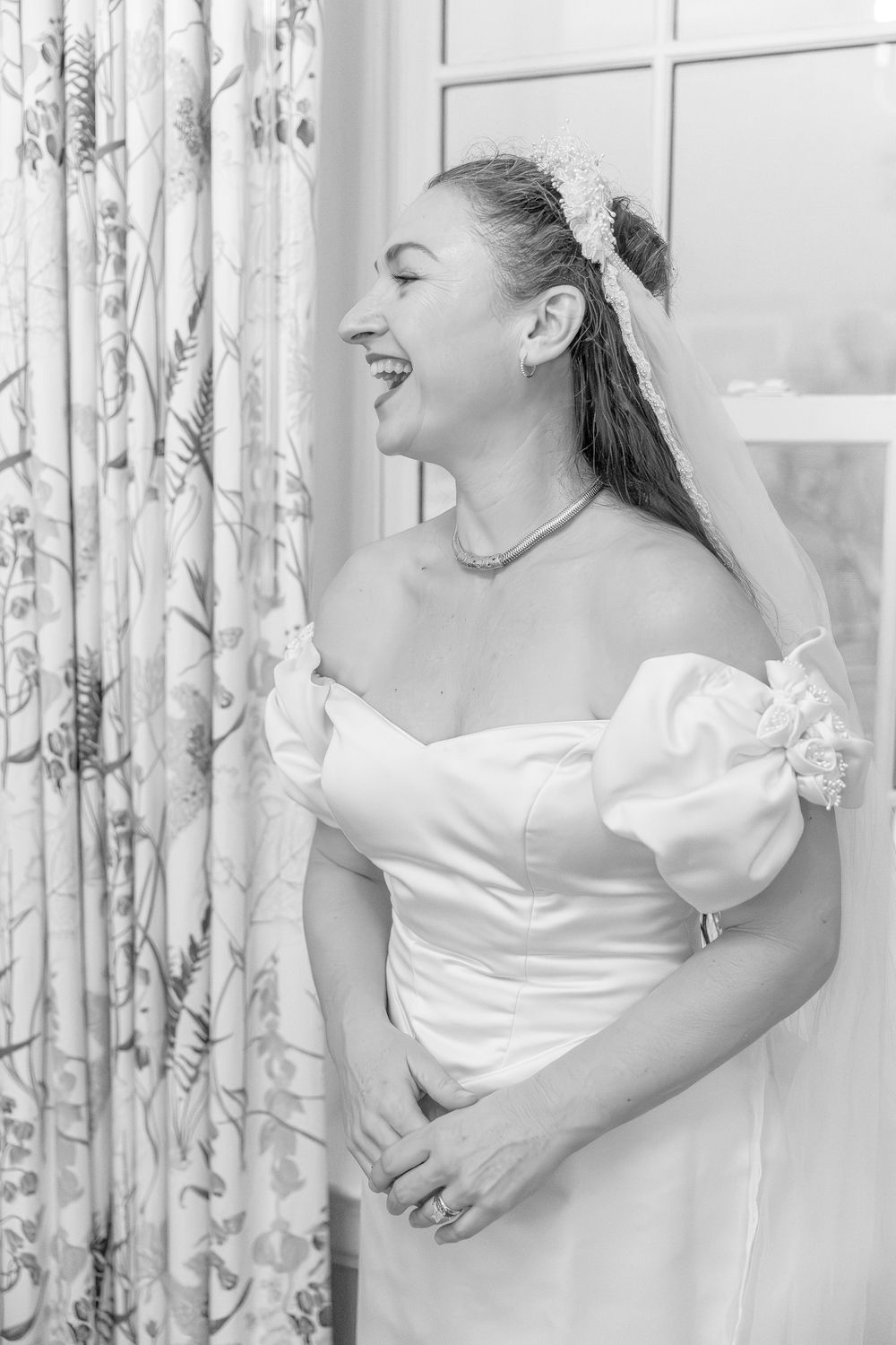 Joyful bride in off-shoulder white gown laughing beside floral curtain, black and white portrait.
