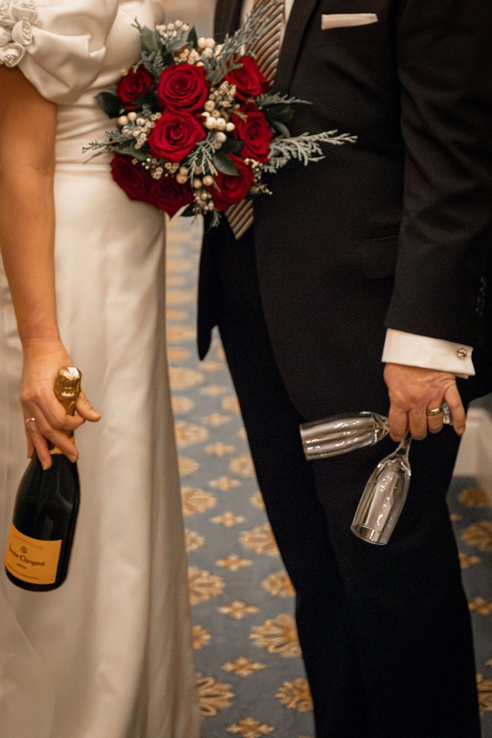 Bride and groom holding Veuve Clicquot champagne bottle and glasses with red rose bridal bouquet.