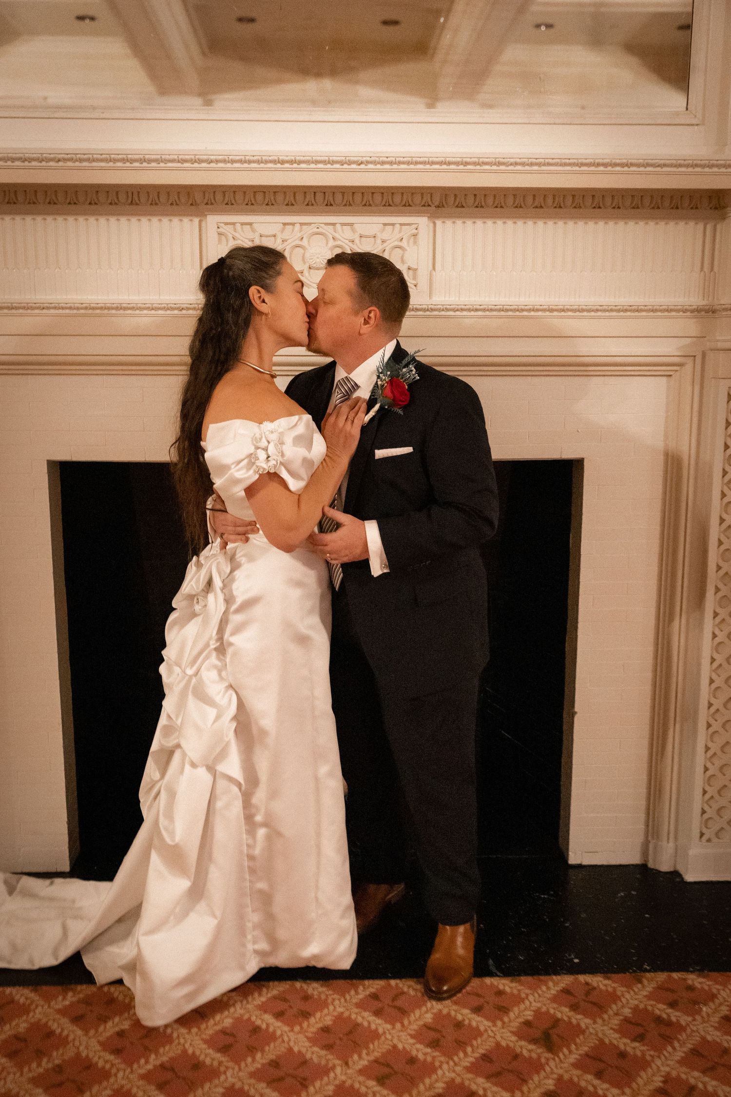 Bride and groom sharing a romantic kiss in front of elegant fireplace during wedding reception.