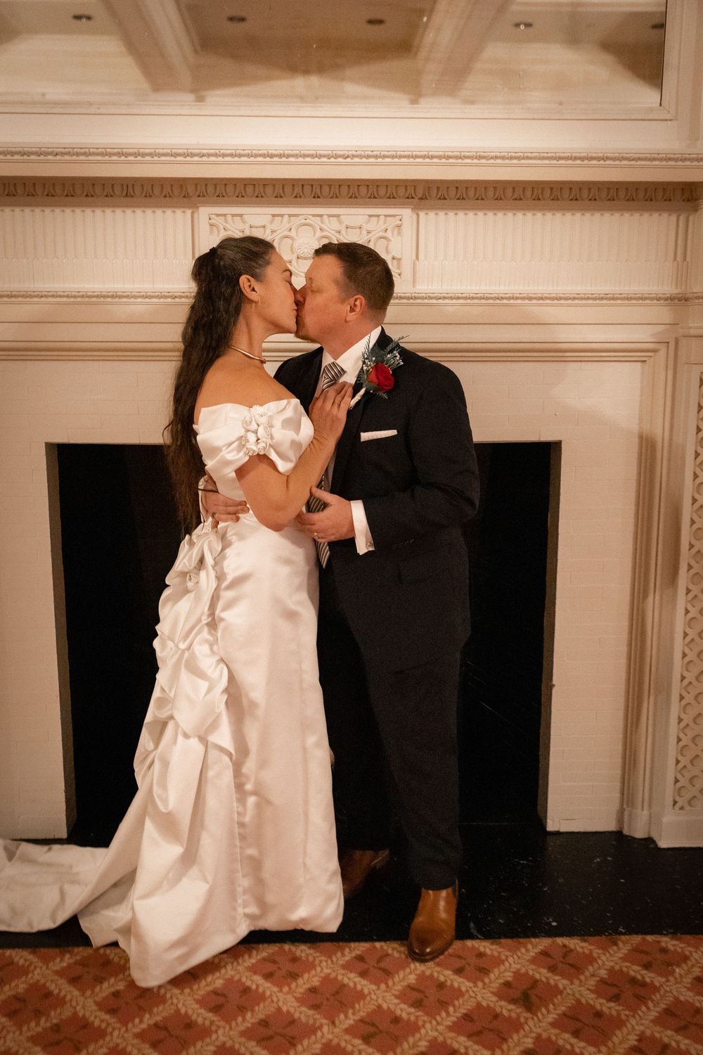Bride and groom sharing a romantic kiss in front of elegant fireplace during wedding reception.