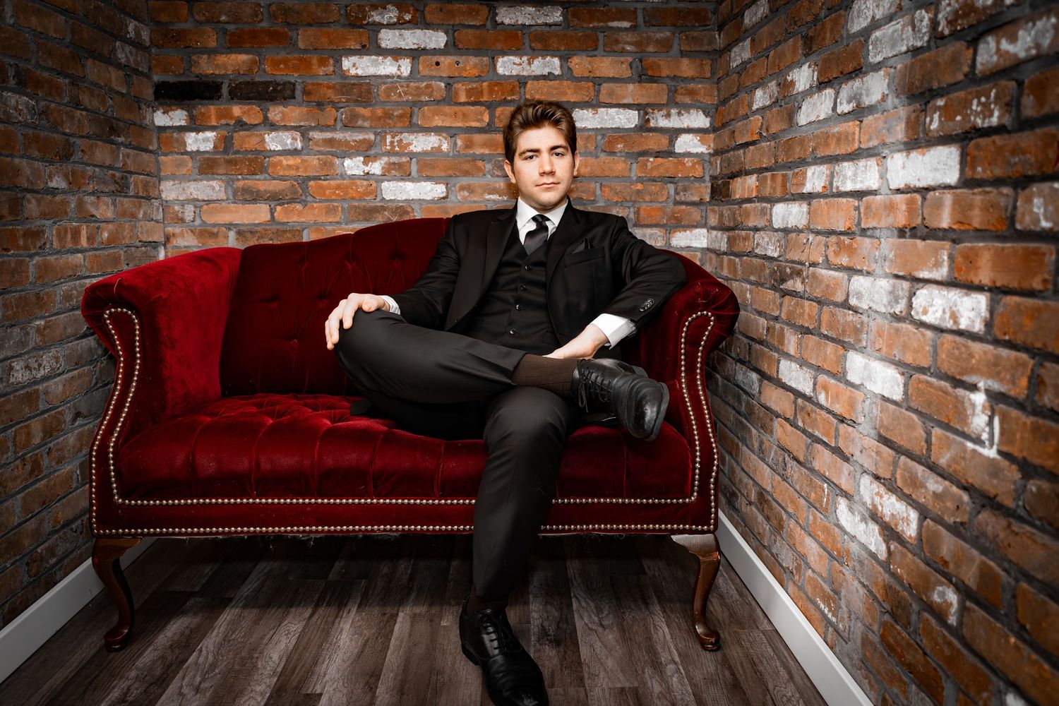 Stylish young man in a black suit sitting on a red velvet sofa against a brick wall.