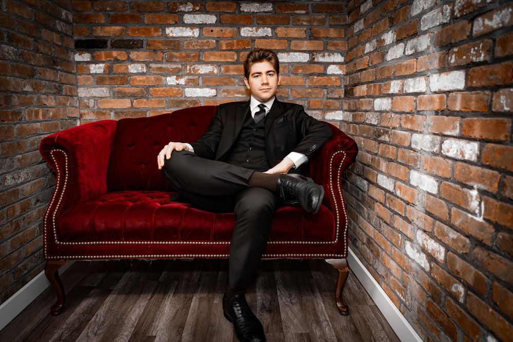 Stylish young man in a black suit sitting on a red velvet sofa against a brick wall.