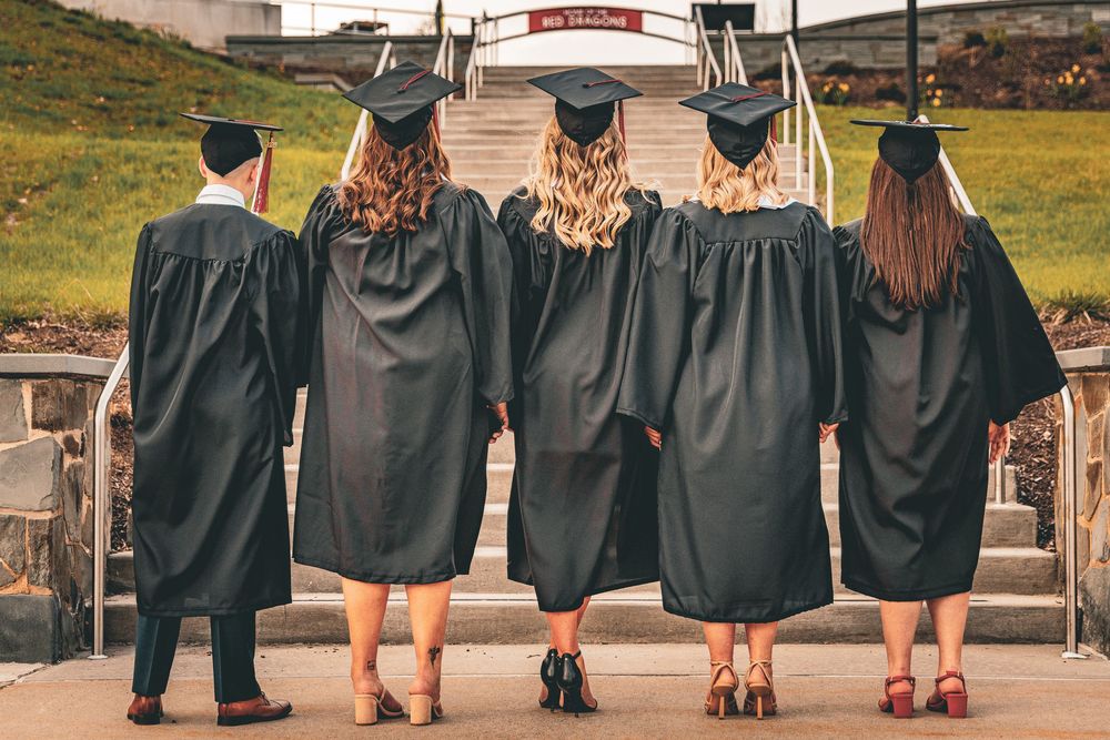 Five graduates in black caps and gowns standing together showing their backs on graduation day.
