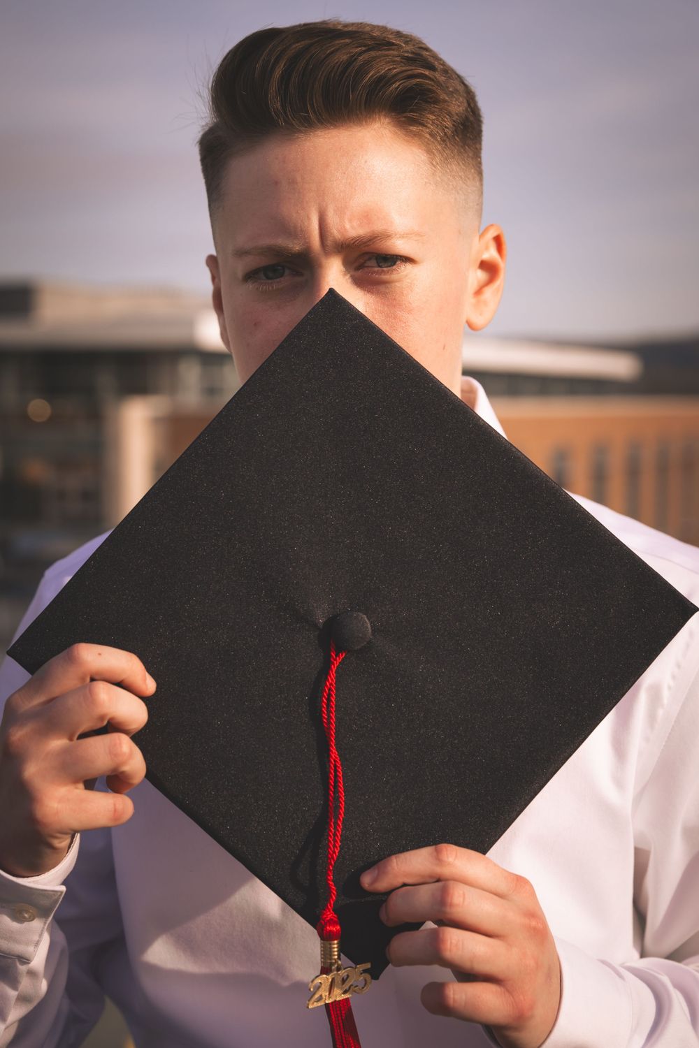 Graduate holding black mortarboard cap with red tassel in front of face outdoors.